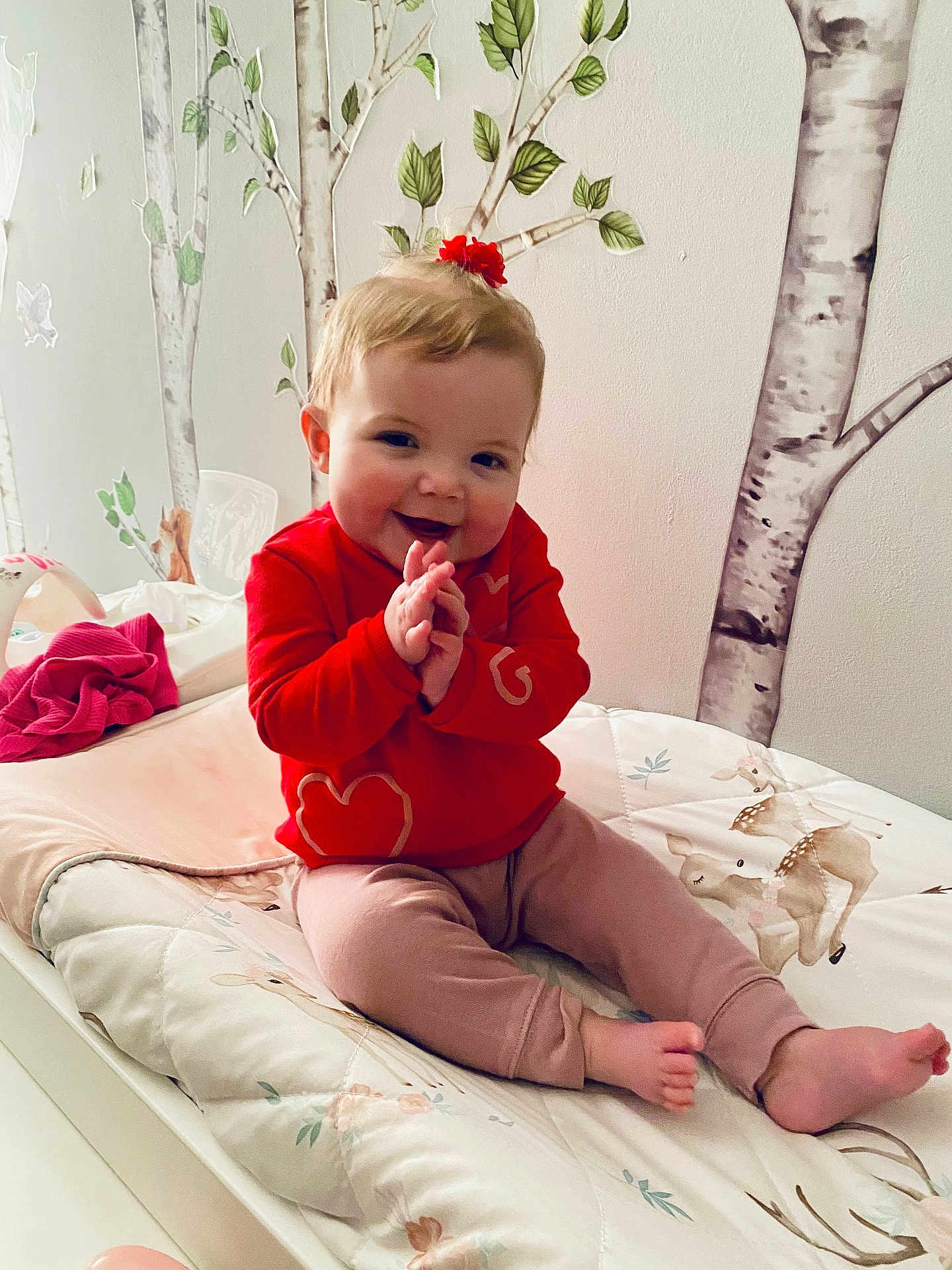 Mila joined the competition — help win amazing prizes! child, baby, toddler, smiling, red_shirt, hair_bow, bed, changing_pad, blanket, wall_mural, birch_trees, nursery, decor, hands_clapping, sitting, toes, pants, indoor, cute, portrait