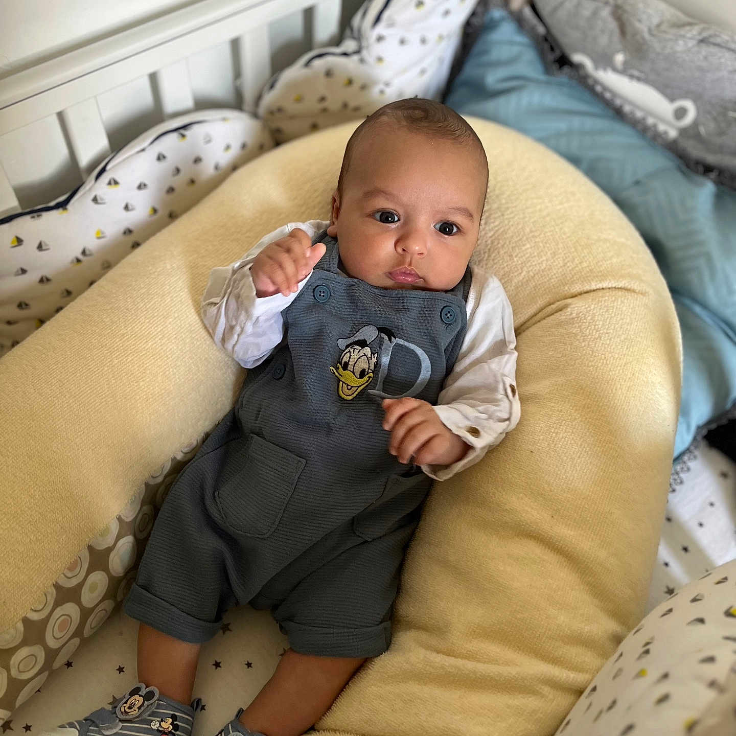 Mohamed Oussaid a rejoint le concours — aidez-le/la à gagner de superbes lots ! baby, baby_nest, child, clothing, comfortable, crib, cushion, cute, cute_outfit, donald_duck, footwear, indoors, infant, mickey_mouse, patterned_bedding, person, resting, sneakers, soft, white_wall