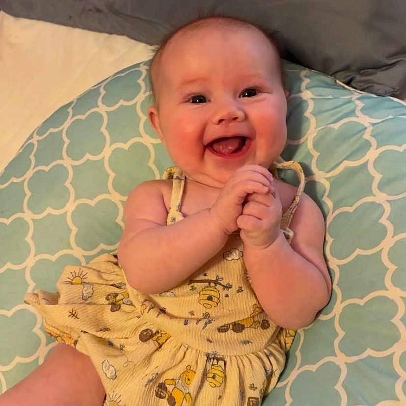 Charlotte is registered to the contest to win money with this photo: baby, smiling, happy, infant, clapping, yellow_dress, patterned_cushion, cute, cheerful, indoors, child, skin, face, arms, hands, joyful, seated, plush, clothing, expression