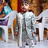 child, toddler, boy, portrait, traditional_clothing, formalwear, wedding, celebration, standing, looking_up, serious_expression, blue_shoes, ornate_outfit, scarf, indoor, decorations, plastic_chairs, guests, colorful_background, photography