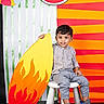 Advait is registered to the contest to win money with this photo: child, boy, toddler, portrait, sitting, stool, smiling, happy, indoors, colorful, backdrop, watermelon, cardboard, flame, prop, striped_background, checkered_shirt, gray_pants, sneakers, photography