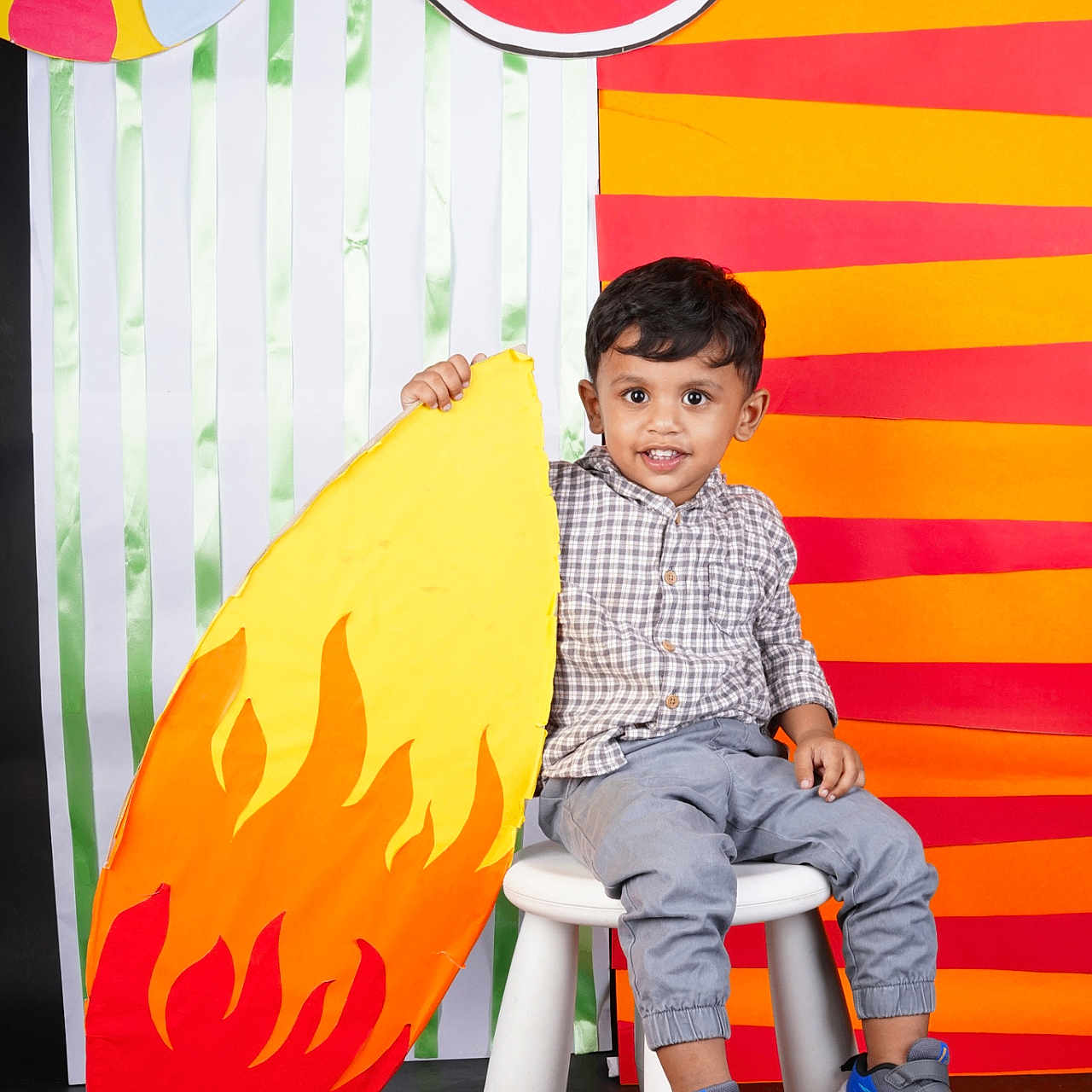 Advait is registered to the contest to win money with this photo: child, boy, toddler, portrait, sitting, stool, smiling, happy, indoors, colorful, backdrop, watermelon, cardboard, flame, prop, striped_background, checkered_shirt, gray_pants, sneakers, photography