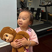 Emmy is registered to the contest to win money with this photo: toddler, child, plush_toy, monkey, indoor, smile, pink_shirt, bracelet, hairclip, toddler_boy, toy, floor, furniture, treadmill, dining_table, chair, portrait, happy, cute, person