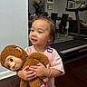 toddler, child, plush_toy, monkey, indoor, smile, pink_shirt, bracelet, hairclip, toddler_boy, toy, floor, furniture, treadmill, dining_table, chair, portrait, happy, cute, person