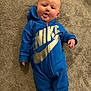 baby, child, blue_onesie, nike_logo, carpet, tongue_out, cute, infant, smiling, lying_down, clothing, footwear, socks, indoors, person, happy, face, head, playful, cozy
