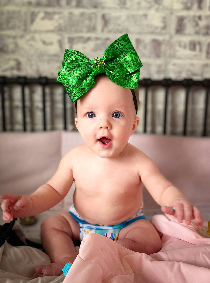 Willow is registered to the contest to win money with this photo: baby, baby_toddler_clothing, child, costume_hat, event, face, fashion_accessory, foot, fun, green, happy, head, headband, headpiece, headwear, human_leg, person, sitting, skin, surprise