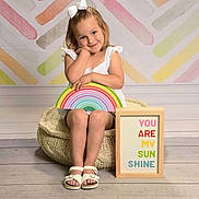 Jenna joined the competition — help win amazing prizes! child, girl, white_bow, rainbow, pouf, sign, colorful, pastel_background, sitting, smiling, white_sandals, floor, wooden_floor, cute, portrait, indoor, happy, decor, message, playful