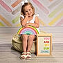 child, girl, white_bow, rainbow, pouf, sign, colorful, pastel_background, sitting, smiling, white_sandals, floor, wooden_floor, cute, portrait, indoor, happy, decor, message, playful