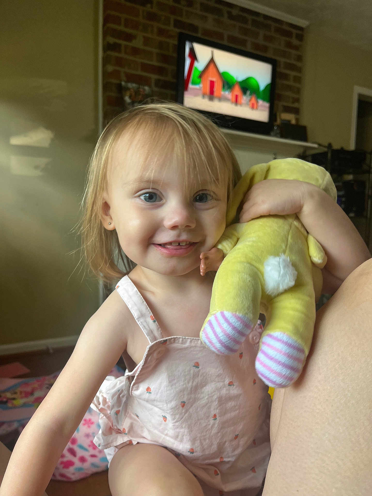Riley is registered to the contest to win money with this photo: toddler, child, smiling, indoor, stuffed_toy, yellow_toy, blue_eyes, light_hair, pink_clothing, sleeveless_outfit, brick_fireplace, television, cartoon, living_room, happy, cute, person, playful, home, portrait