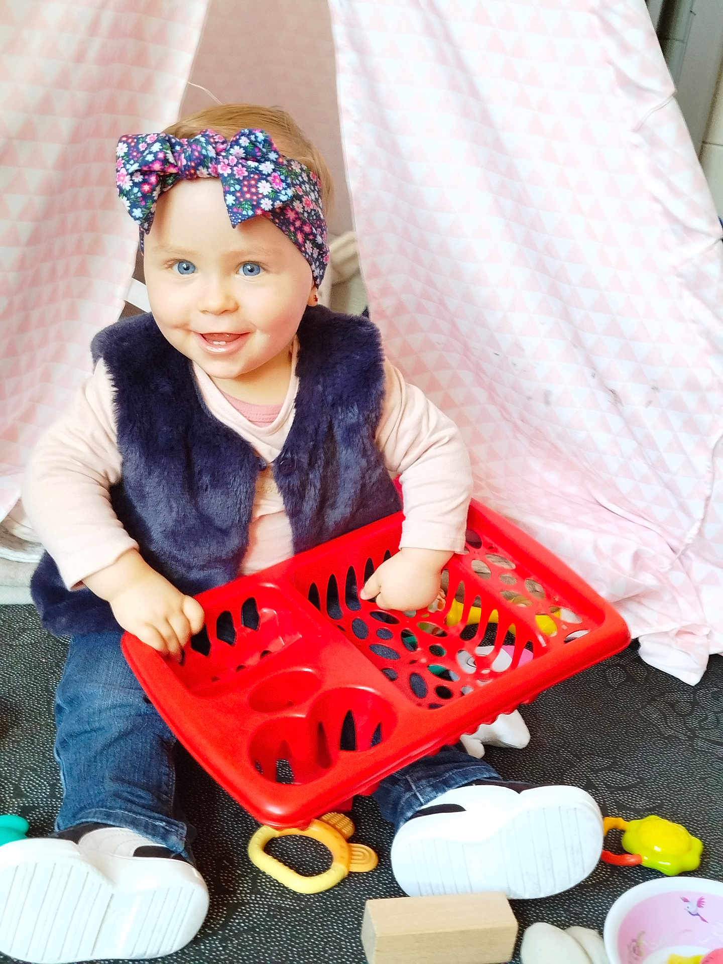 Anna a rejoint le concours — aidez-le/la à gagner de superbes lots ! toddler, child, blue_eyes, headband, floral, red_basket, toys, sitting, smiling, indoor, cute, happy, casual_clothing, jeans, white_shoes, playtime, floor, pink_backdrop, vest, candid
