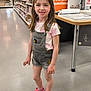child, girl, smile, standing, denim_overalls, pink_clogs, sunglasses, store_aisle, floor, display_table, shelves, clothing, shorts, casual_wear, retail, happy, indoor, person, footwear, young