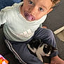 toddler, child, pacifier, curly_hair, blue_eyes, kitten, cat, couch, clothing, sweatshirt, pants, hand, foot, smartphone, indoor, cozy, pet, sitting, looking_up, casual