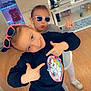 child, kids, indoor, sunglasses, unicorn, sweatshirt, playing, pose, peace_sign, floor, wooden_floor, tv_screen, video_game, toys, shelf, casual_clothing, smile, fun, home, person