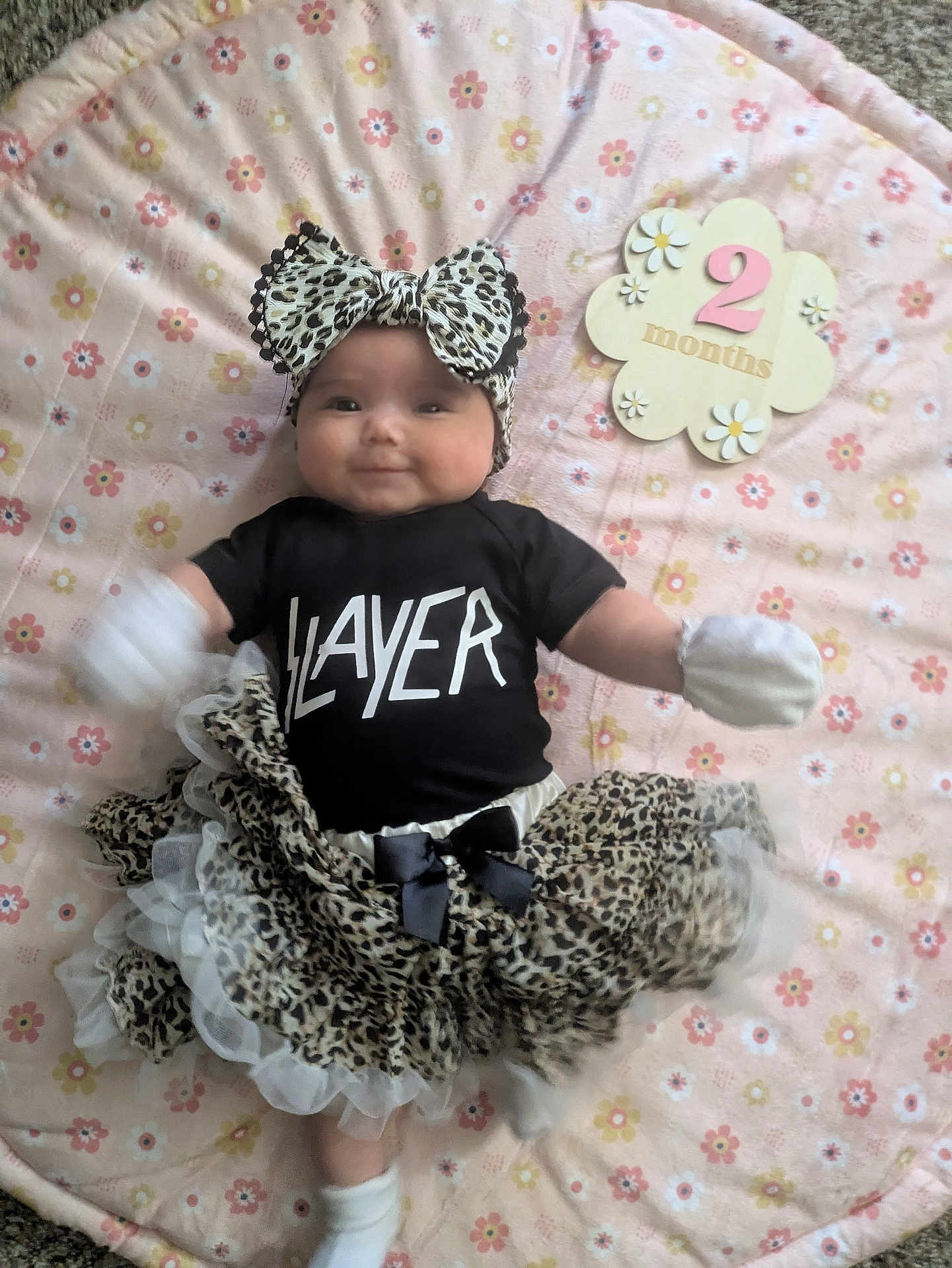 Allie is registered to the contest to win money with this photo: baby, infant, tutu, headband, black_shirt, slayer, leopard_print, mittens, socks, blanket, floral_pattern, wooden_sign, 2_months, smiling, lying_down, cute, child, clothing, person, indoor