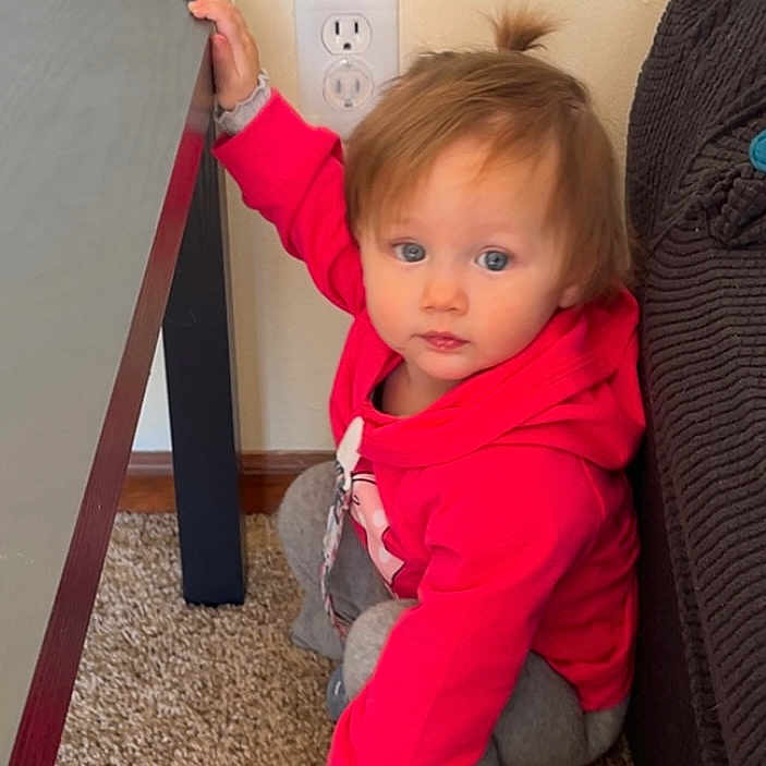 Belladonna June is registered to the contest to win money with this photo: baby, blue_eyes, brown_hair, carpet, casual_clothing, child, couch, cute, hand_on_table, hoodie, indoor, living_room, outlet, pony_tail, red_hoodie, sitting, squatting, table, toddler, wall
