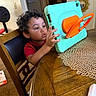 child, curly_hair, tablet, protective_case, wooden_table, placemat, chair, home, indoor, person, technology, curiosity, young, sitting, furniture, red_shirt, carpet, doorway, mirror, hand