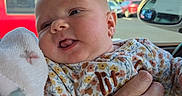 Oakleigh is registered to the contest to win money with this photo: baby, infant, child, face, smiling, tongue_out, portrait, held, adult_hand, floral_clothing, blanket, car, vehicle, car_interior, outdoor, blue_sky, trees, parked_cars, daylight, cozy