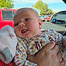 baby, infant, child, face, smiling, tongue_out, portrait, held, adult_hand, floral_clothing, blanket, car, vehicle, car_interior, outdoor, blue_sky, trees, parked_cars, daylight, cozy
