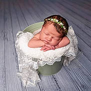 Amari is registered to the contest to win money with this photo: newborn, baby, sleeping, flower_crown, lace, blanket, bucket, wooden_floor, infant, peaceful, cute, portrait, resting, soft, decor, head, hands, skin, young_child, indoors