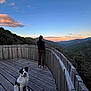 dog, person, mountains, sunset, wooden_deck, sky, clouds, outdoor, nature, happy_dog, landscape, scenic_view, trees, fence, evening, animal, pet, walking_shoes, leisure, relaxation