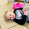 child, boy, smiling, lying_down, floor, tile_floor, blonde_hair, nike, sweatshirt, long_sleeve, indoor, toy, pink_toy, background_blur, happy, casual_clothing, portrait, person, young, cute