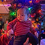 baby, child, person, indoor, christmas_wreath, holiday_decorations, ornaments, colorful_lights, red_shirt, striped_clothing, hand_holding, tattoo, festive, celebration, warm_lighting, curtains, gift_boxes, furniture, cozy, portrait