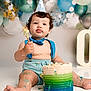baby, cake, birthday, party_hat, blue_suspender, messy, cake_smash, balloons, celebration, one_year_old, child, sitting, food, smile, fun, colorful, cute, indoor, festive, portrait