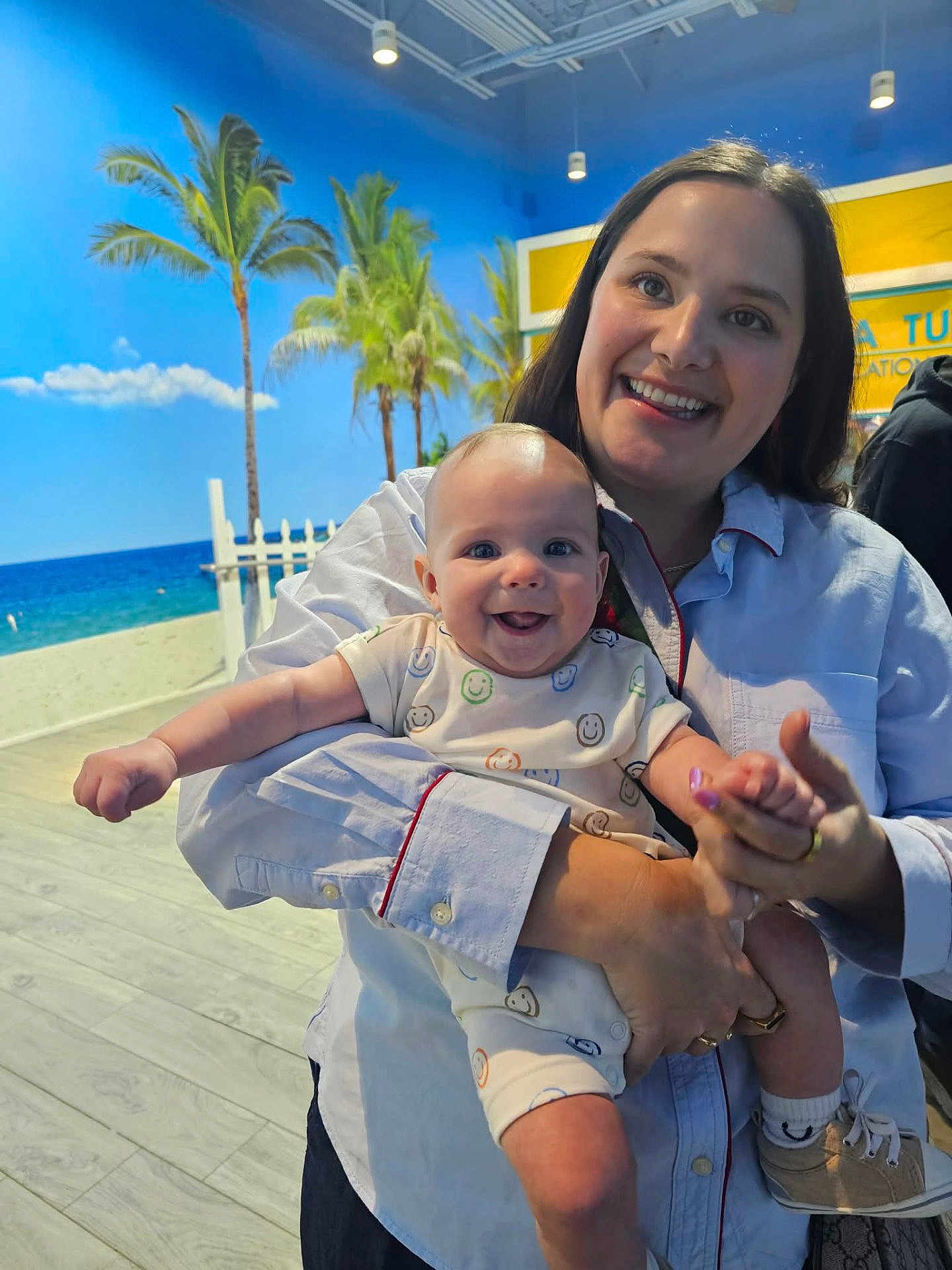 Atticus is registered to the contest to win money with this photo: baby, woman, smiling, holding, indoor, tropical_mural, palm_trees, blue_sky, beach_scene, onesie, happy, person, portrait, clothing, flooring, hand, footwear, light, background, expression