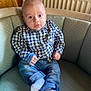 baby, child, infant, plaid_shirt, jeans, socks, couch, furniture, indoors, radiator, wood_paneling, face, person, seated, curious, casual_clothing, home, portrait, young_child, comfortable