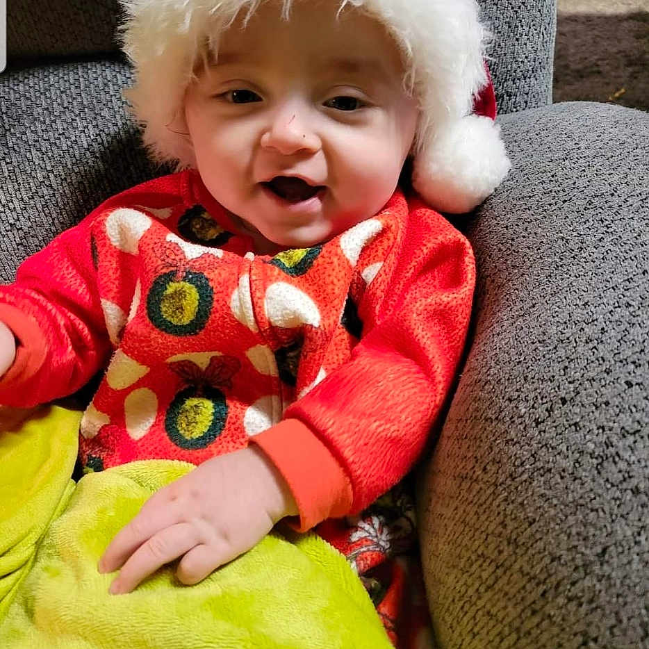 Major is registered to the contest to win money with this photo: baby, child, infant, portrait, smiling, happy, santa_hat, christmas, festive, red_clothing, green_blanket, chair, indoor, seated, face, eyes, mouth, photograph, cozy, holiday