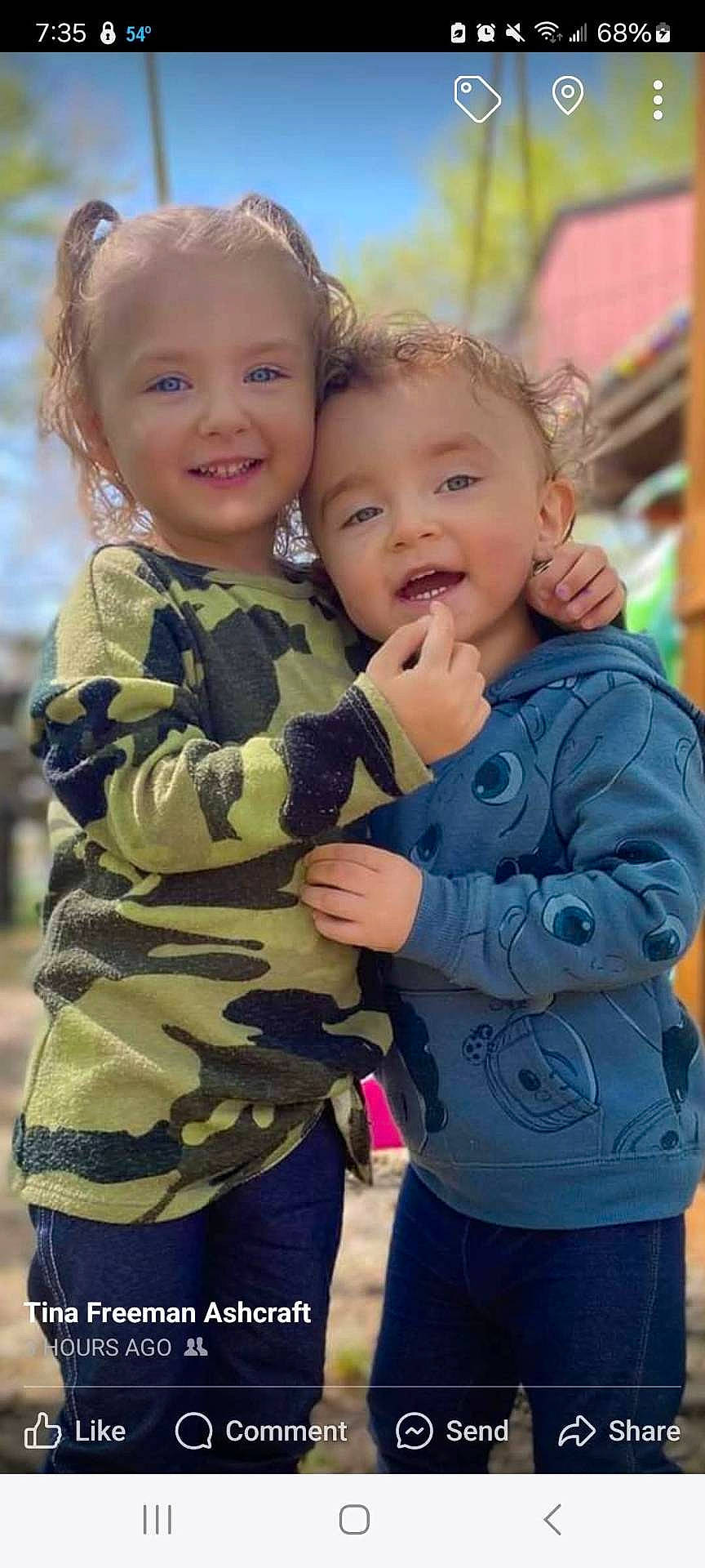 blue_hoodie, blue_sky, building, camouflage_clothing, children, curly_hair, daylight, greenery, happy, hugging, jeans, outdoor, phone_screenshot, playful, portrait, preschooler, siblings, smiling, social_media, toddler