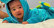 Niklaus is registered to the contest to win money with this photo: baby, child, dinosaur_costume, dinosaur_hood, turquoise, blue, smiling, happy, portrait, indoors, bed, bedding, blanket, white_bedding, lying_down, costume, hood, infant, face, playful