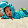 baby, child, dinosaur_costume, dinosaur_hood, turquoise, blue, smiling, happy, portrait, indoors, bed, bedding, blanket, white_bedding, lying_down, costume, hood, infant, face, playful