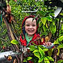 child, smiling, hooded_jacket, green_leaves, bush, outdoor, nature, happy, face, person, plants, trees, young, curious, playful, red, green, daylight, joyful, portrait