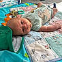baby, infant, quilt, blanket, toy, clothing, hat, dinosaur_hat, lying_down, blue, green, cute, small_person, indoor, child, face, expression, soft_toys, floor, patterned