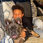 Chanel is registered to the contest to win money with this photo: child, dog, puppy, couch, pillow, blanket, smiling, happy, indoors, cozy, curly_hair, black_shirt, light_skin, ears, pet, face, furniture, home, relaxing, playful