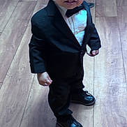 Kingstyn is registered to the contest to win money with this photo: toddler, child, suit, bow_tie, curly_hair, black_shoes, wooden_floor, indoor, formal_wear, standing, person, flooring, furniture, chair, legs, smile, cute, fashion, portrait, young