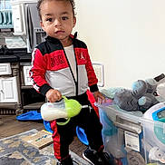 Kashtyn is registered to the contest to win money with this photo: toddler, child, indoor, toy_bin, sippy_cup, plush_elephant, rug, kitchen, clothing, sneakers, person, floor, cabinet, wall, photograph, standing, curly_hair, expression, home, playtime