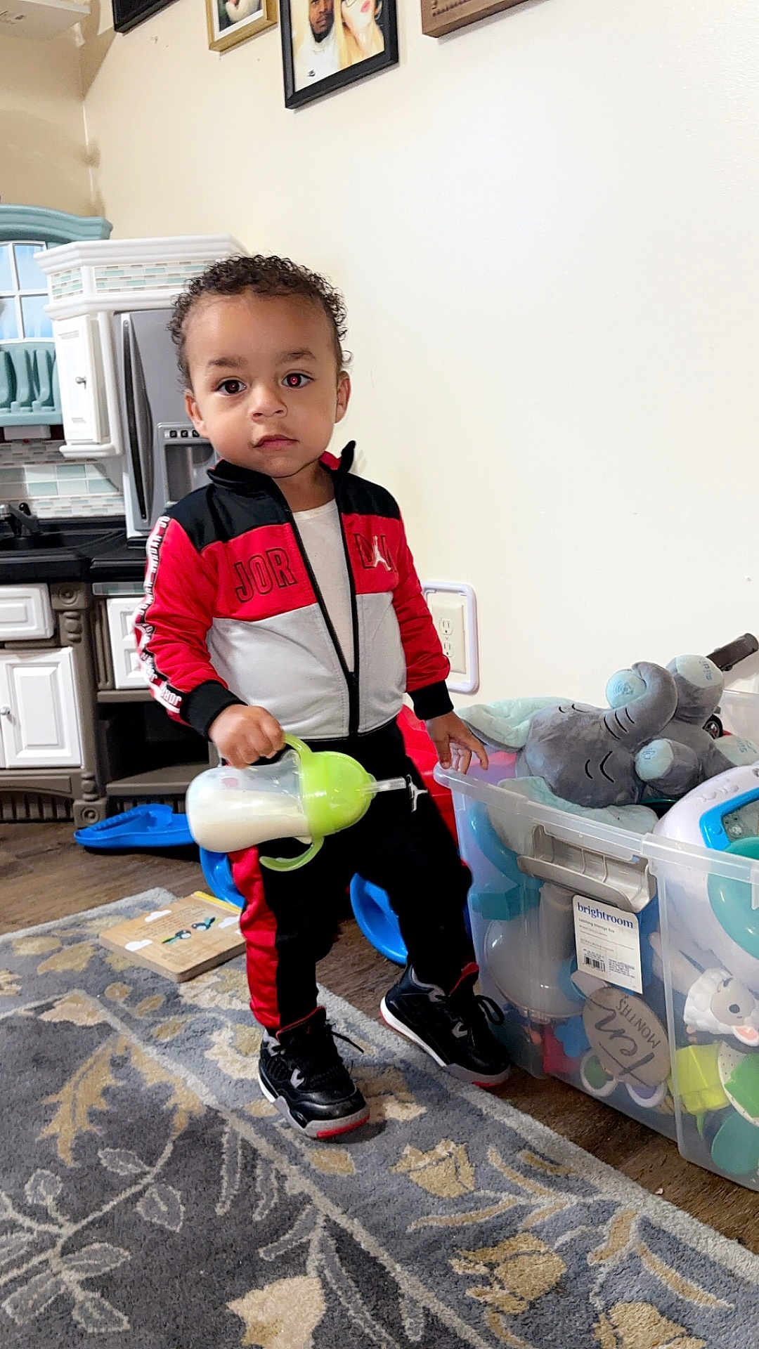 Kashtyn is registered to the contest to win money with this photo: toddler, child, indoor, toy_bin, sippy_cup, plush_elephant, rug, kitchen, clothing, sneakers, person, floor, cabinet, wall, photograph, standing, curly_hair, expression, home, playtime