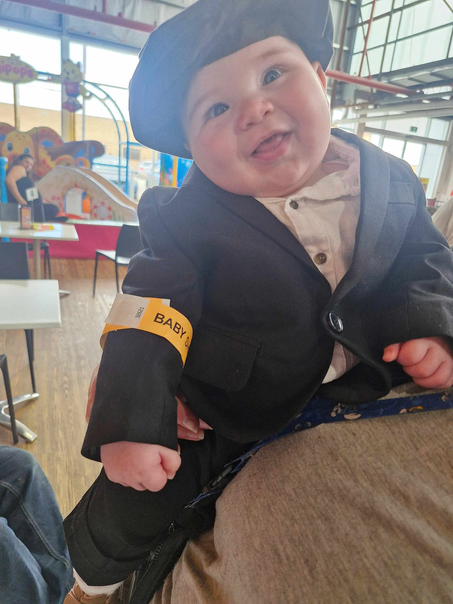 Walter joined the competition — help win amazing prizes! baby, child, smiling, face, hat, suit, jacket, wristband, indoor, play_area, person, people, seated, table, chair, wood_floor, window, light, happy, cute