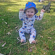 Walter is registered to the contest to win money with this photo: baby, child, grass, outdoor, hat, bucket_hat, onesie, cartoon_print, smiling, happy, sitting, park, daylight, playful, barefoot, clothing, nature, person, greenery, sunlight