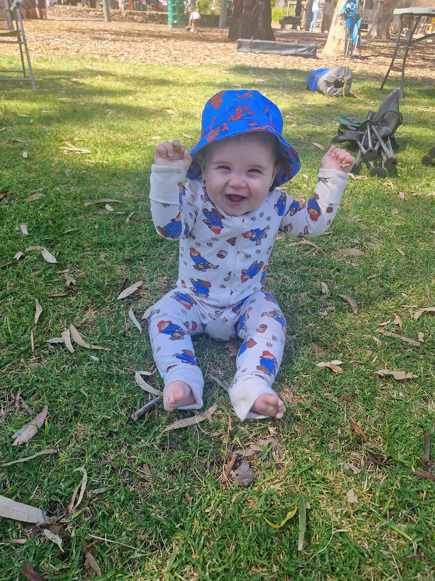 Walter is registered to the contest to win money with this photo: baby, child, grass, outdoor, hat, bucket_hat, onesie, cartoon_print, smiling, happy, sitting, park, daylight, playful, barefoot, clothing, nature, person, greenery, sunlight
