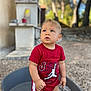 toddler, child, red_clothing, jordan_logo, outdoor, daylight, trees, gravel_ground, gray_tub, curious_expression, standing, shorts, short_sleeves, blond_hair, face, person, nature, sunlight, blurred_background, young_child