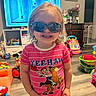 Barbie Rae is registered to the contest to win money with this photo: child, toddler, girl, portrait, smiling, sunglasses, indoors, playroom, toys, television, pink_shirt, striped_clothing, cowboy_theme, cartoon_shirt, standing, playpen, dresser, colorful, family_home, happy