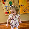 blonde_hair, bow, child, colorful, cute, dress, gears, indoor, minnie_mouse, pacifier, person, playroom, shoes, socks, toddler, toys, walking, wall_toys, wooden_floor, young_child