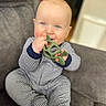 baby, child, teether, toy, blue_eyes, striped_clothing, onesie, couch, gray_couch, indoor, smiling, happy, sitting, infant, cute, chewing, hand, face, skin, young_child