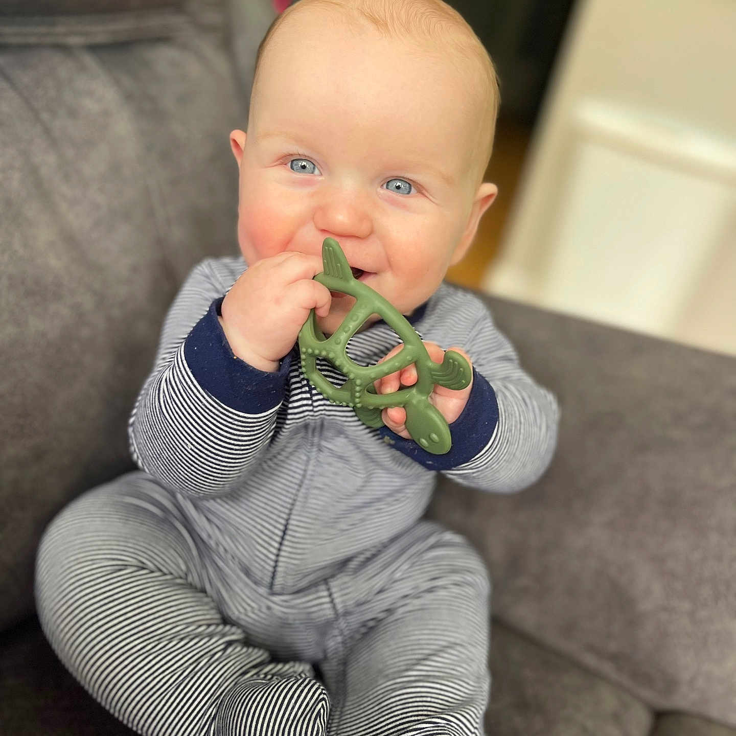 Malaki is registered to the contest to win money with this photo: baby, child, teether, toy, blue_eyes, striped_clothing, onesie, couch, gray_couch, indoor, smiling, happy, sitting, infant, cute, chewing, hand, face, skin, young_child