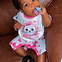 baby, child, pacifier, bow, curly_hair, brown_couch, pink_clothing, cat_illustration, slippers, fuzzy_slippers, cute, indoor, sitting, infant, playful, cozy, portrait, happy, young_child, clothing