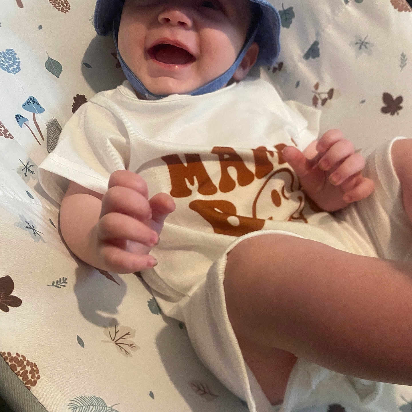 Hunter Jay is registered to the contest to win money with this photo: apparel, baby, blue_hat, child, closeup, cute, face, hand, happy, hat, indoors, infant, leaves_pattern, lying_down, patterned_cushion, pinecone_pattern, playful, smiling, toddler, white_clothing