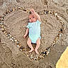 baby, infant, child, beach, sand, pebbles, heart_shape, outdoor, lying_down, onesie, mint_green, raised_hand, portrait, face, sleepy, calm, sunlight, natural_texture, creative_arrangement, family_photo
